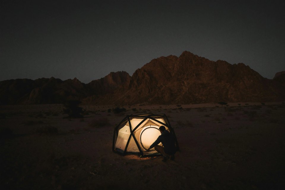 A person setting up a modern tent in a desert landscape at night, with mountains in the background and a warm glow emanating from the tent.