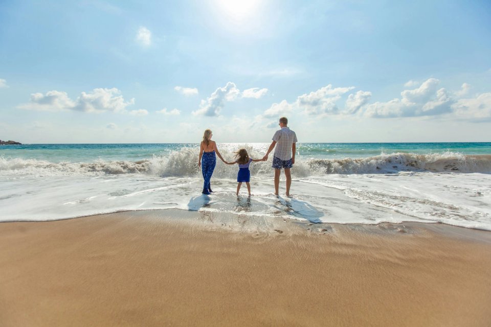 A family of three, including a woman, a girl, and a man, standing hand in hand at the beach, facing the ocean waves under a bright blue sky.