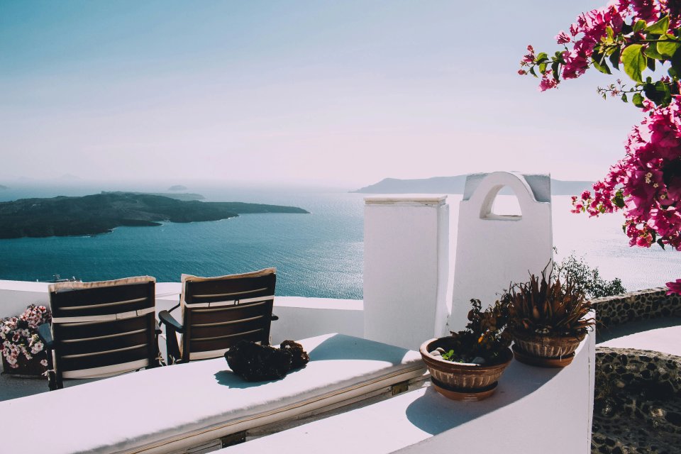 A serene seaside view from Santorini, featuring two chairs overlooking the blue waters, with vibrant flowers and a white architectural structure in the foreground.