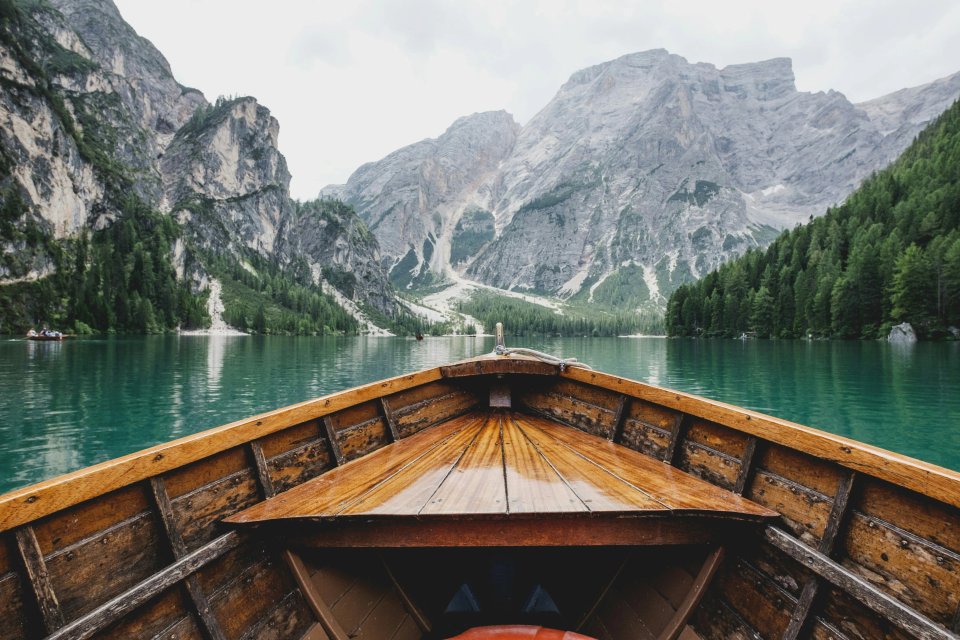 A wooden boat's bow facing a tranquil lake surrounded by towering mountains and lush green trees.