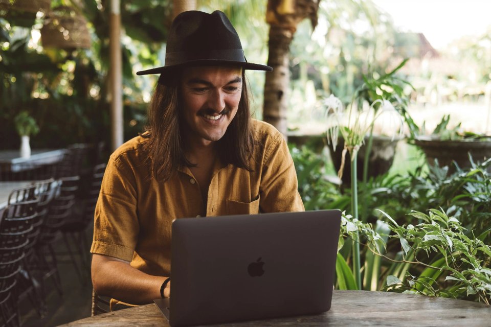 A smiling man with long hair and a hat working on a laptop in a lush outdoor setting surrounded by greenery.