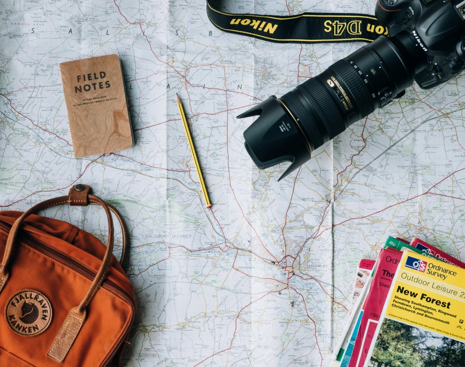A flat lay of travel essentials including a camera, a map, a notebook, a pencil, and a backpack, showcasing preparation for an outdoor adventure.