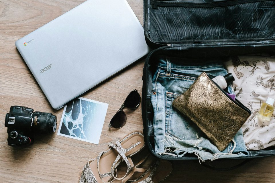 A flat lay of travel essentials including a laptop, camera, sunglasses, denim shorts, a gold clutch, and a photo, arranged on a wooden surface.